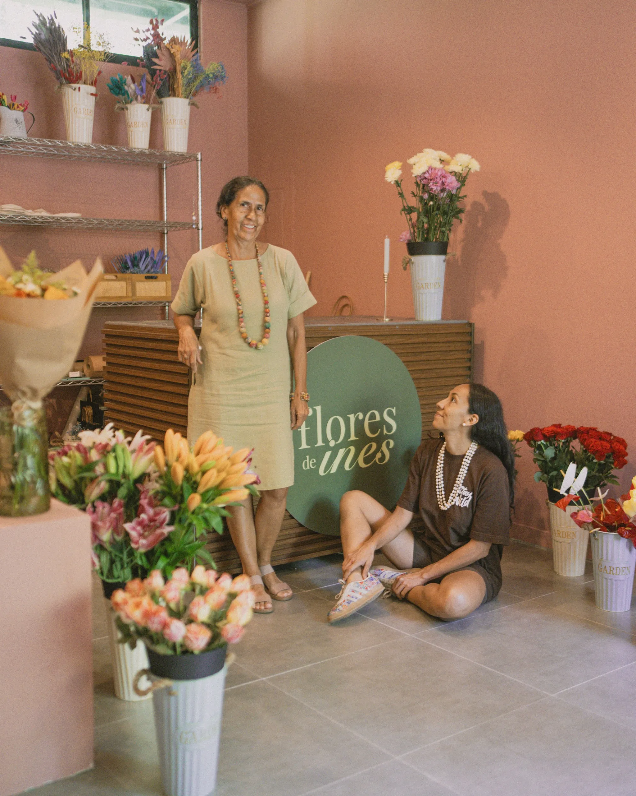 Two women inside Flores de Ines flower shop, surrounded by colorful bouquets and floral arrangements on shelves and buckets