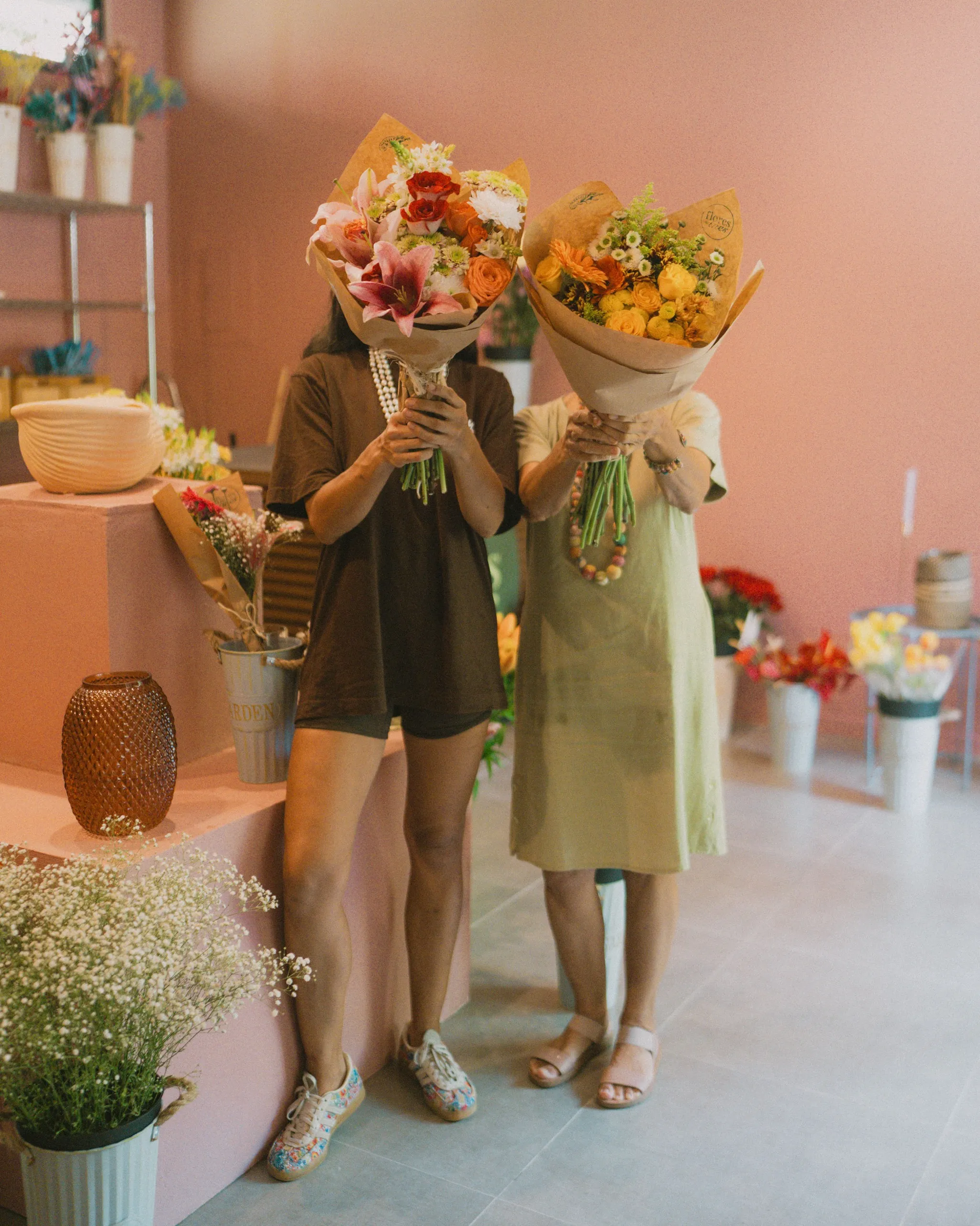 Two people holding large colorful bouquets in front of their faces inside a flower shop with pink walls