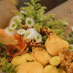Close-up of a floral bouquet featuring peach roses, orange gerbera daisies, white button mums, and yellow chrysanthemums with green foliage
