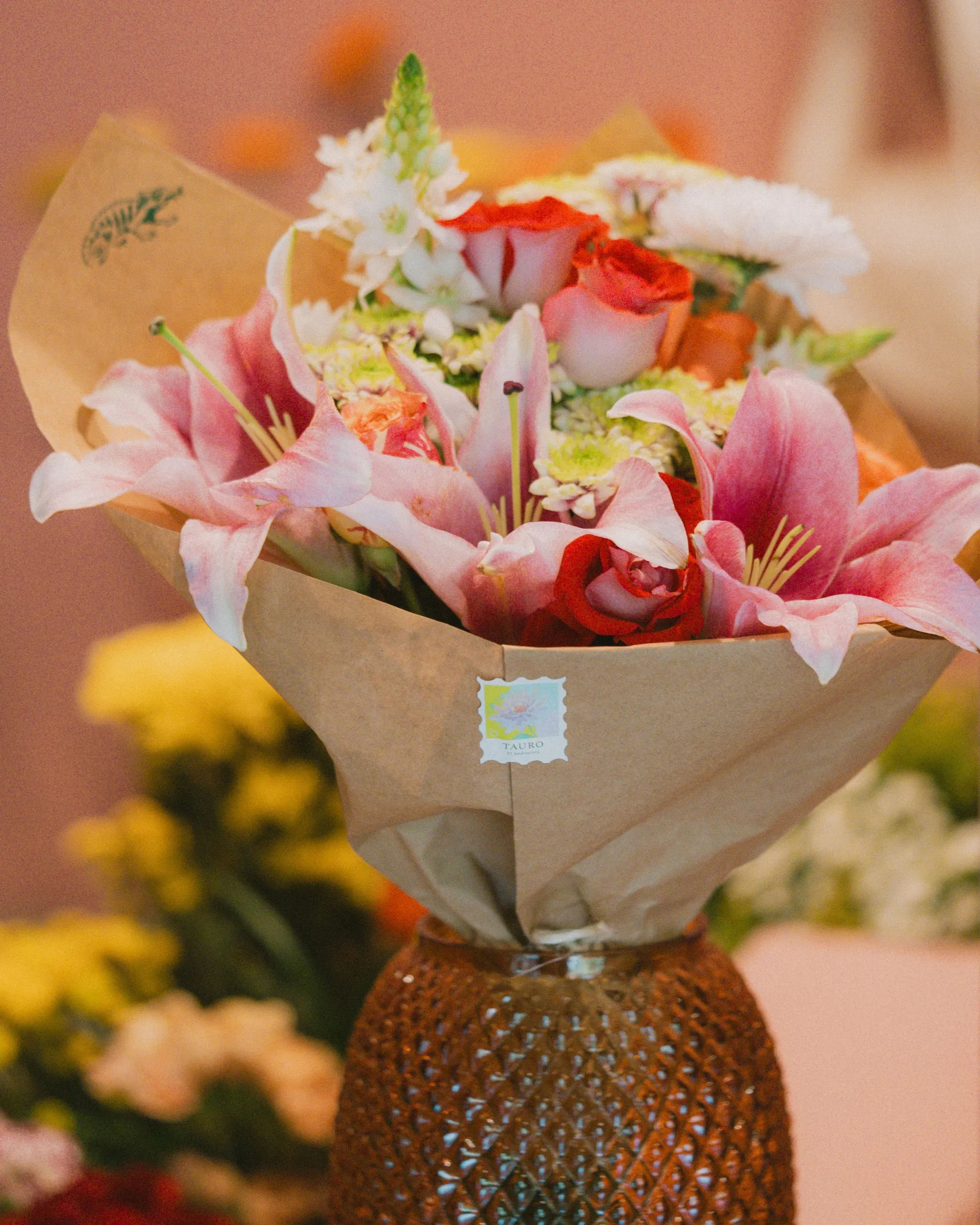 Tauro-branded bouquet of pink lilies, red roses, and white flowers wrapped in kraft paper, displayed in a textured vase