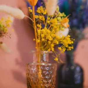 Small amber textured glass vase holding yellow dried flowers and bunny tail grass, with additional floral arrangements in the background