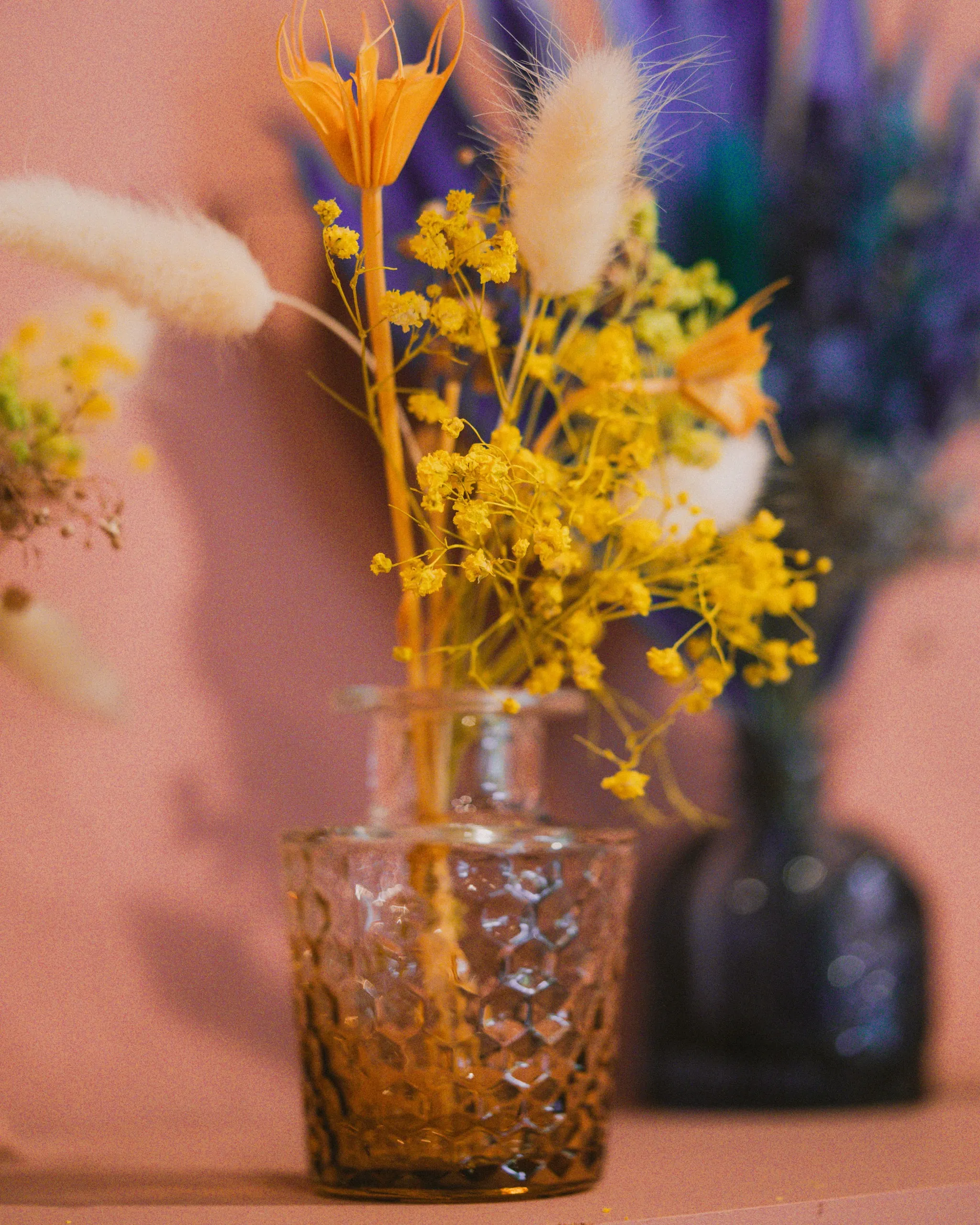 Small amber textured glass vase holding yellow dried flowers and bunny tail grass, with additional floral arrangements in the background