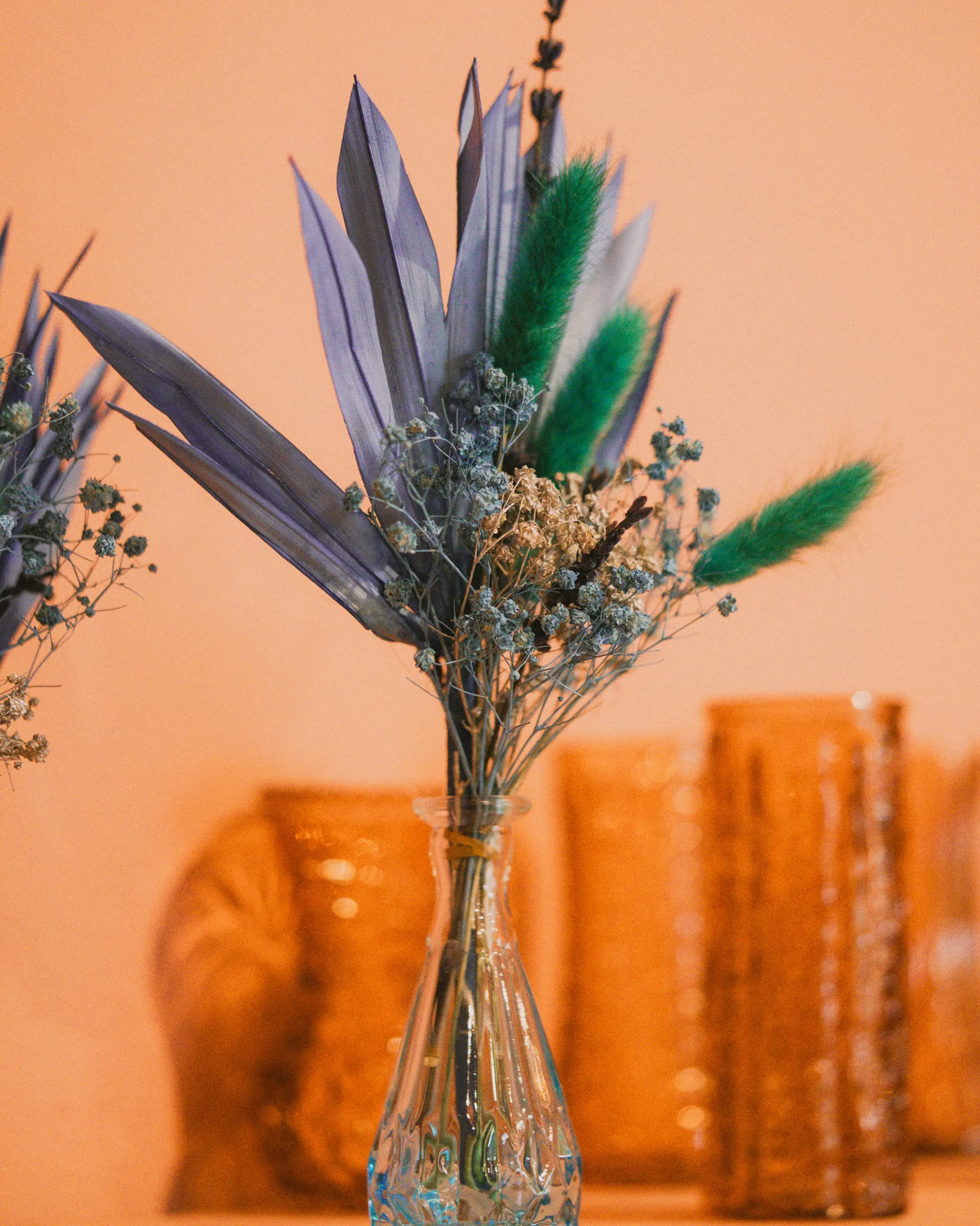 Dried floral arrangement with blue-purple leaves and green fluffy stems in a clear glass vase against a peach background