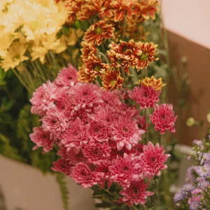 Bunches of pink, orange-yellow, and cream chrysanthemums with purple wildflowers arranged in a white box