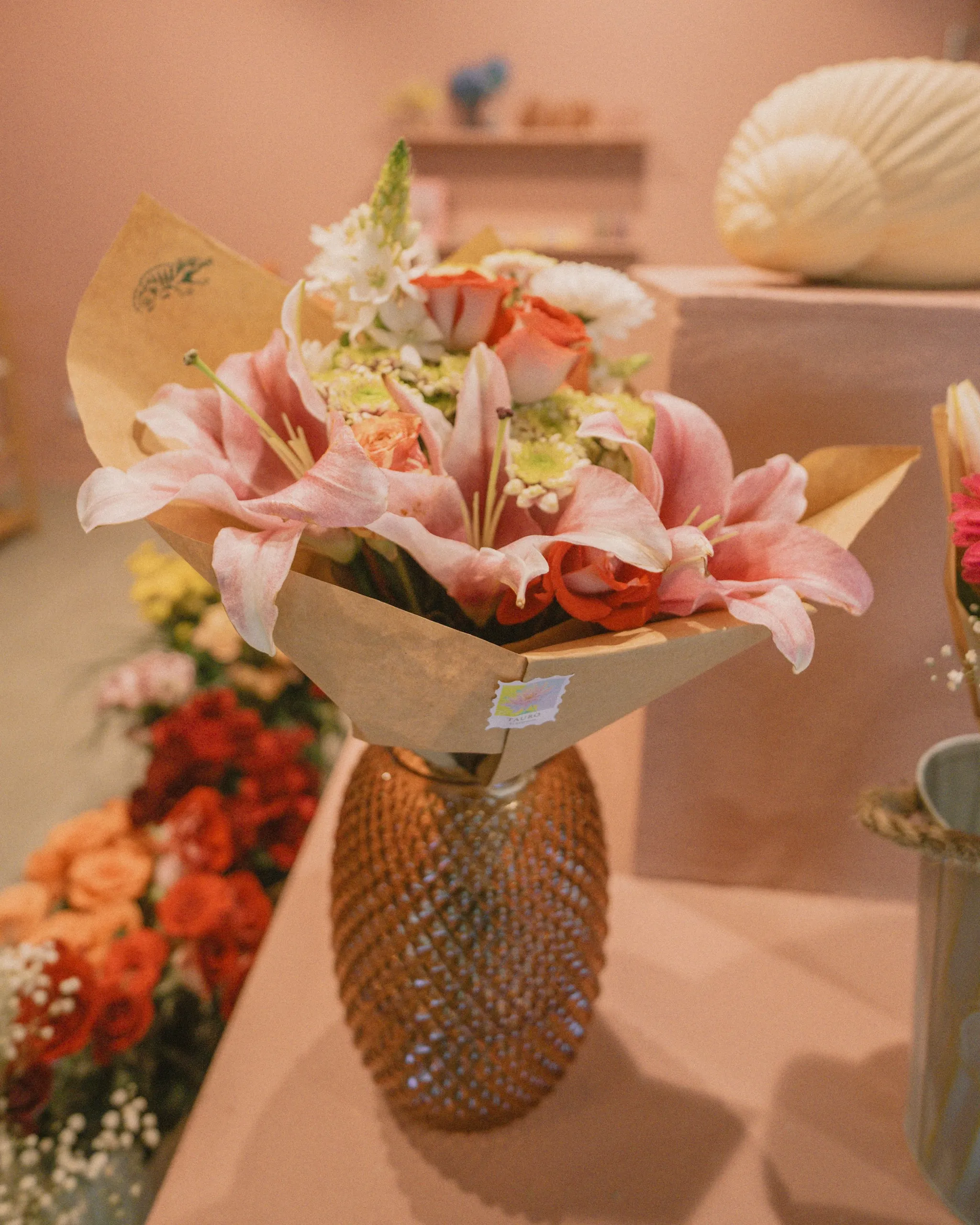 Bouquet of pink lilies, orange roses, and white flowers wrapped in kraft paper, displayed in a textured golden vase inside a flower shop