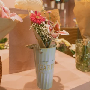 Light blue metal garden bucket holding a mixed bouquet of pink gerbera daisies, baby's breath, and lilies on a table