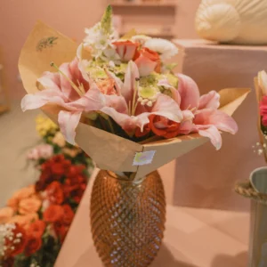 Bouquet of pink lilies and orange roses wrapped in kraft paper, displayed in a textured golden vase inside a flower shop